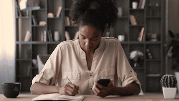 woman writing down a list in her notebook while checking her phone