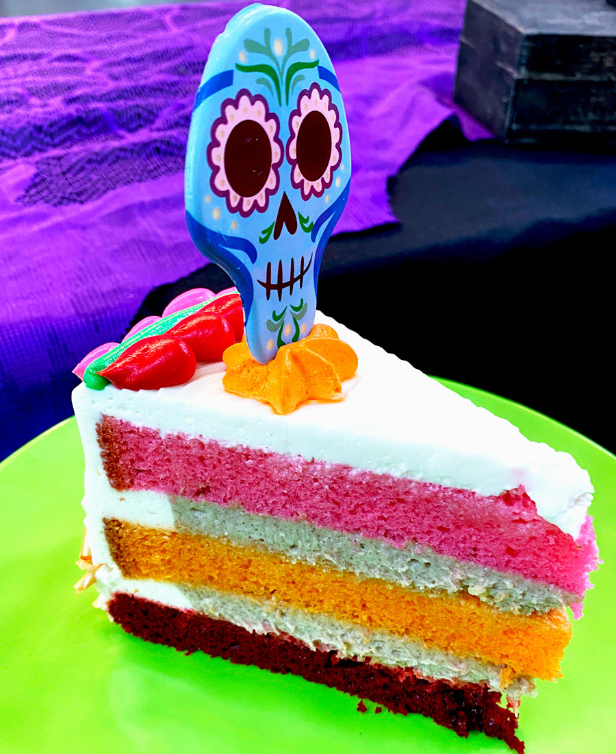 A three layered cake with 3 layers in pink, orange, and red, and a skull shaped decoration on top that has a cute face with flower eyes that&#x27;s smiling