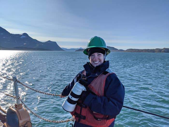 SN Kate Kilroy poses on the bow of a ship. The coast of Greenland is in the background.
