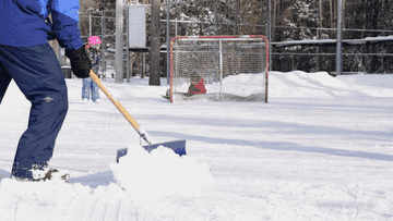 man shoveling snow off an outdoor rink while kids skate in the background