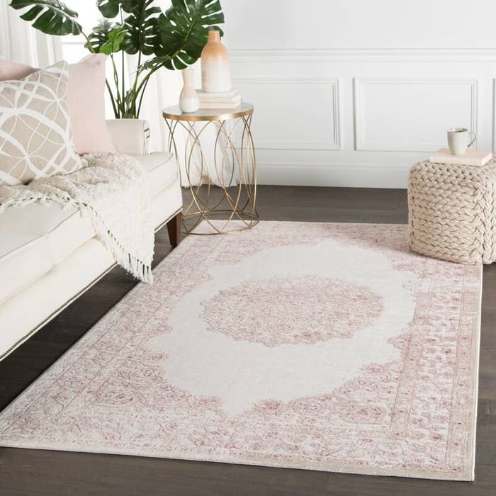 Pink and white medallion area rug placed in a living room with a sofa, end table, floor plant and woven pouf.