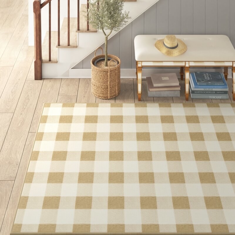 Plaid tan and beige rug in an entryway next to a staircase and a bench and floor plant.