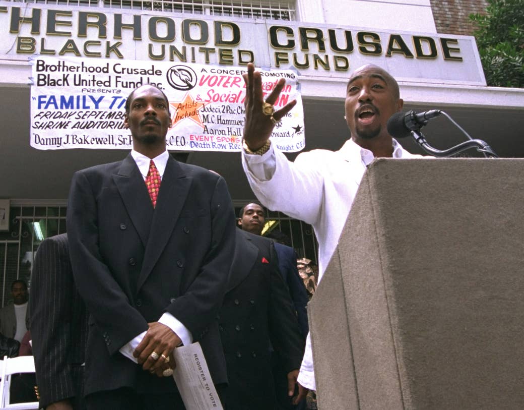 Tupac stands behind a lectern, speaks into a microphone, and gestures with his hand, standing next to Snoop Dogg in a suit