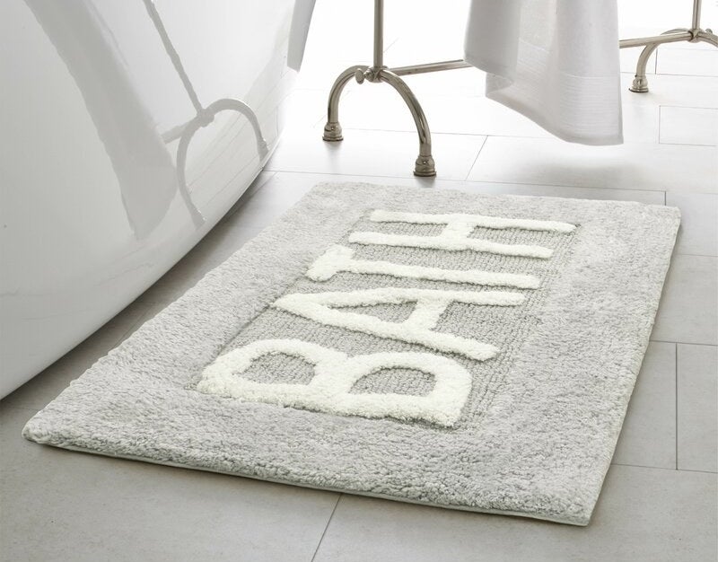Gray rug with the word &quot;BATH&quot; written on it in white, on a tile floor next to a tub.