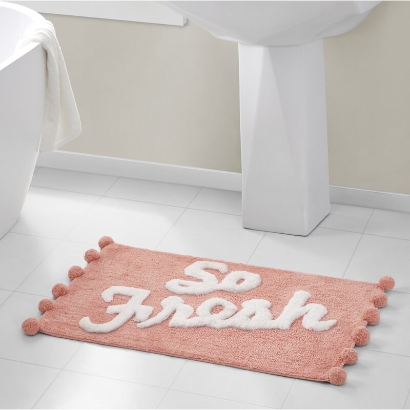 Pink bath rug with the words &quot;So Fresh&quot; written on it in white, on a tile floor in front of a sink.