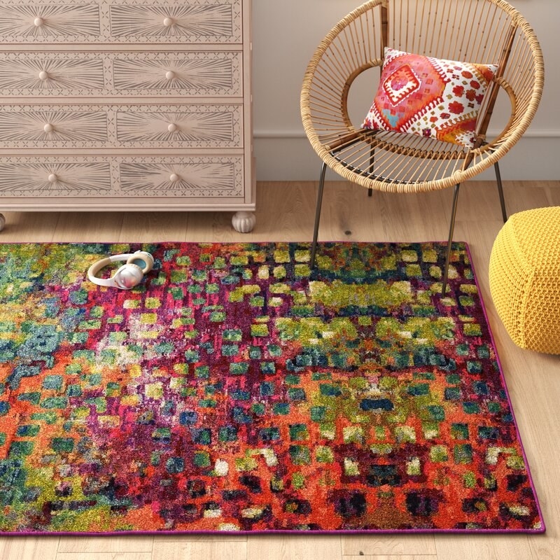 Bright multi-colored rug in a room with a rattan chair, pouf and dresser.