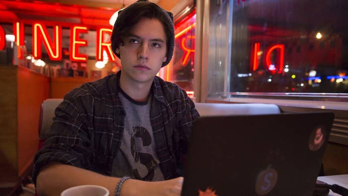 Cole Sprouse as Jughead Jones sitting in a corner booth at a cafe with a laptop on the table