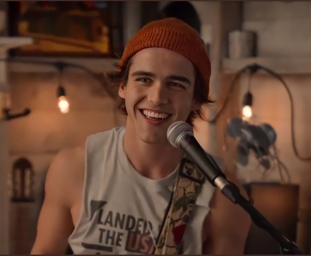 Charlie as Luke in a beanie and vest t-shirt smiling with a microphone in front of him and a guitar strap across his schoulders