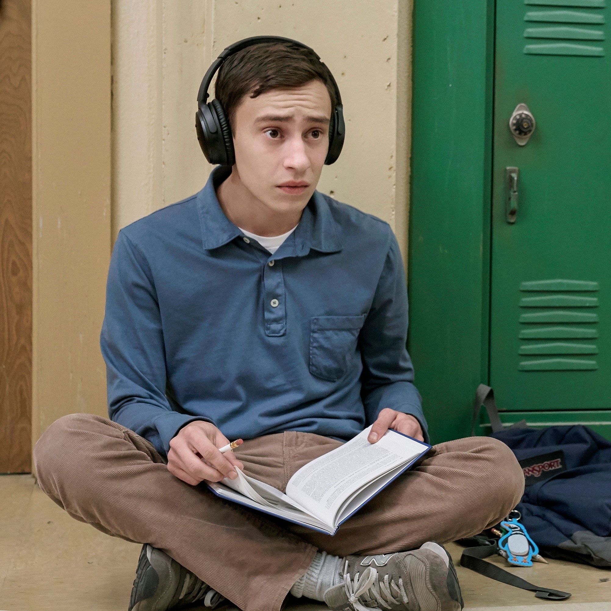 Keir as Sam sitting on the floor with folded legs, holding a book in his lap and a pencil in his hand. He is also wearing headphones