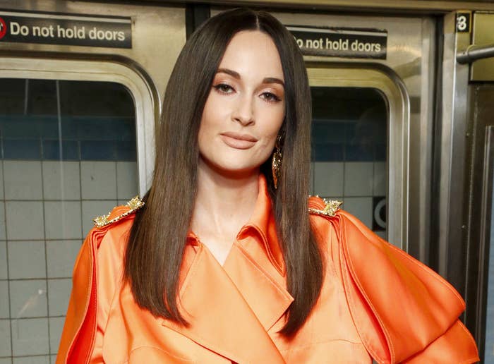 Kacey stands on a subway while wearing a billowing orange dress