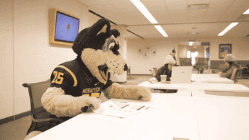 School mascot sits at a table looking at paper before stuffing them into his mouth.