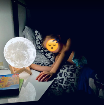 reviewer photo of their child in bed with the moon lamp lit up on the side table