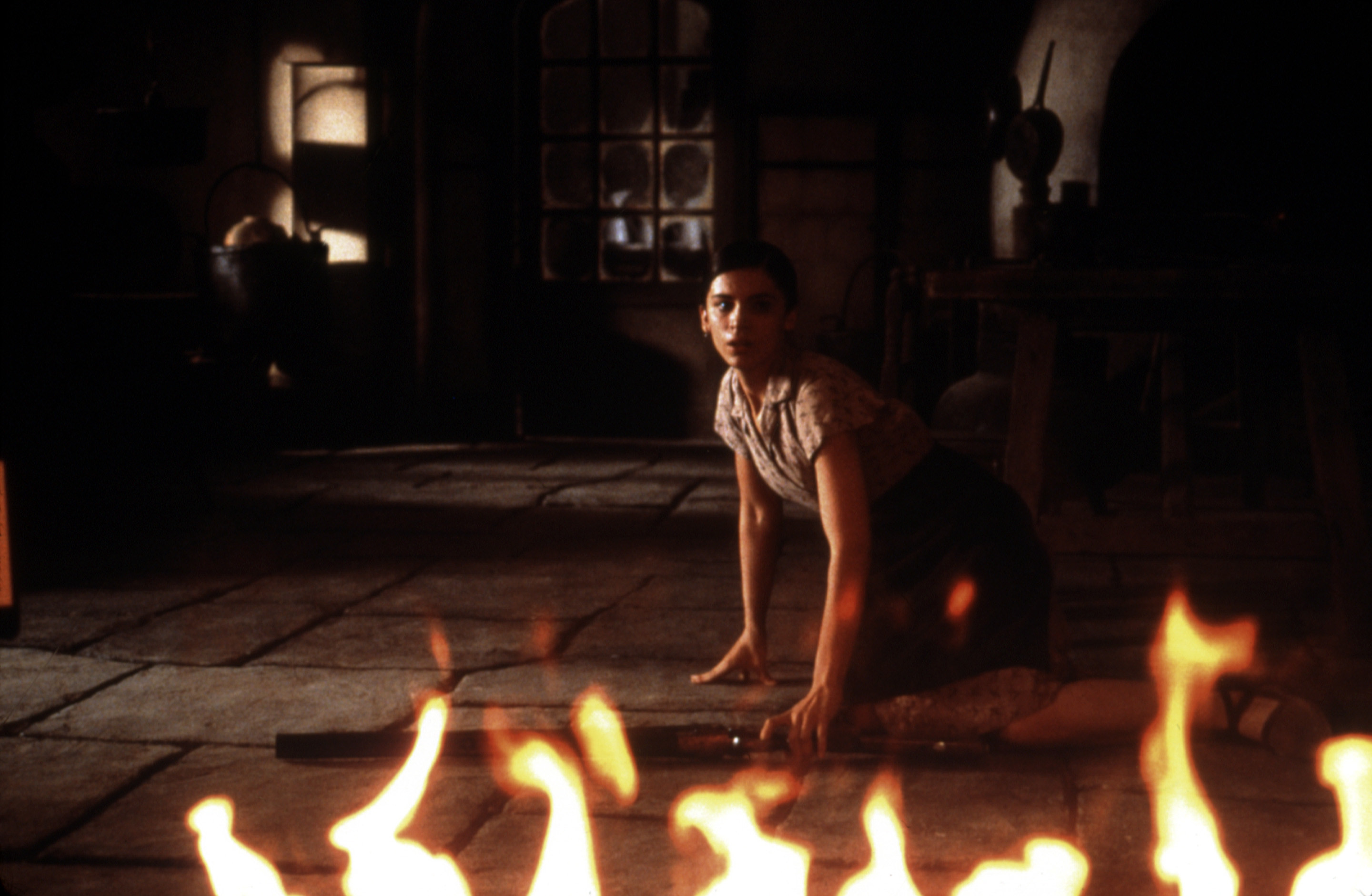 A woman kneeling on the floor, reaching for a rifle as the room goes up in flames around her