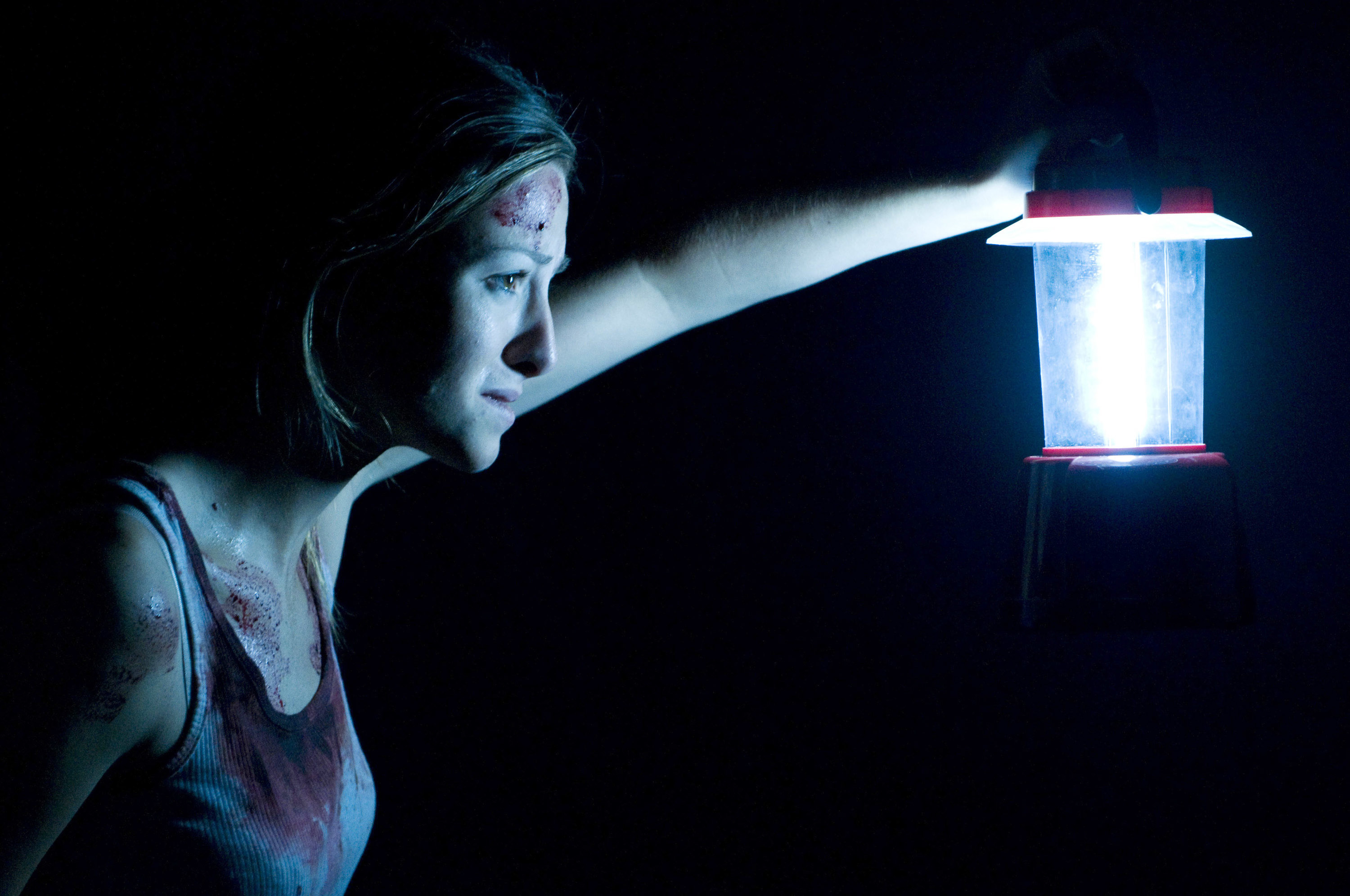 A scared woman, blood on her shirt and face, holding up a lantern in a pitch black room