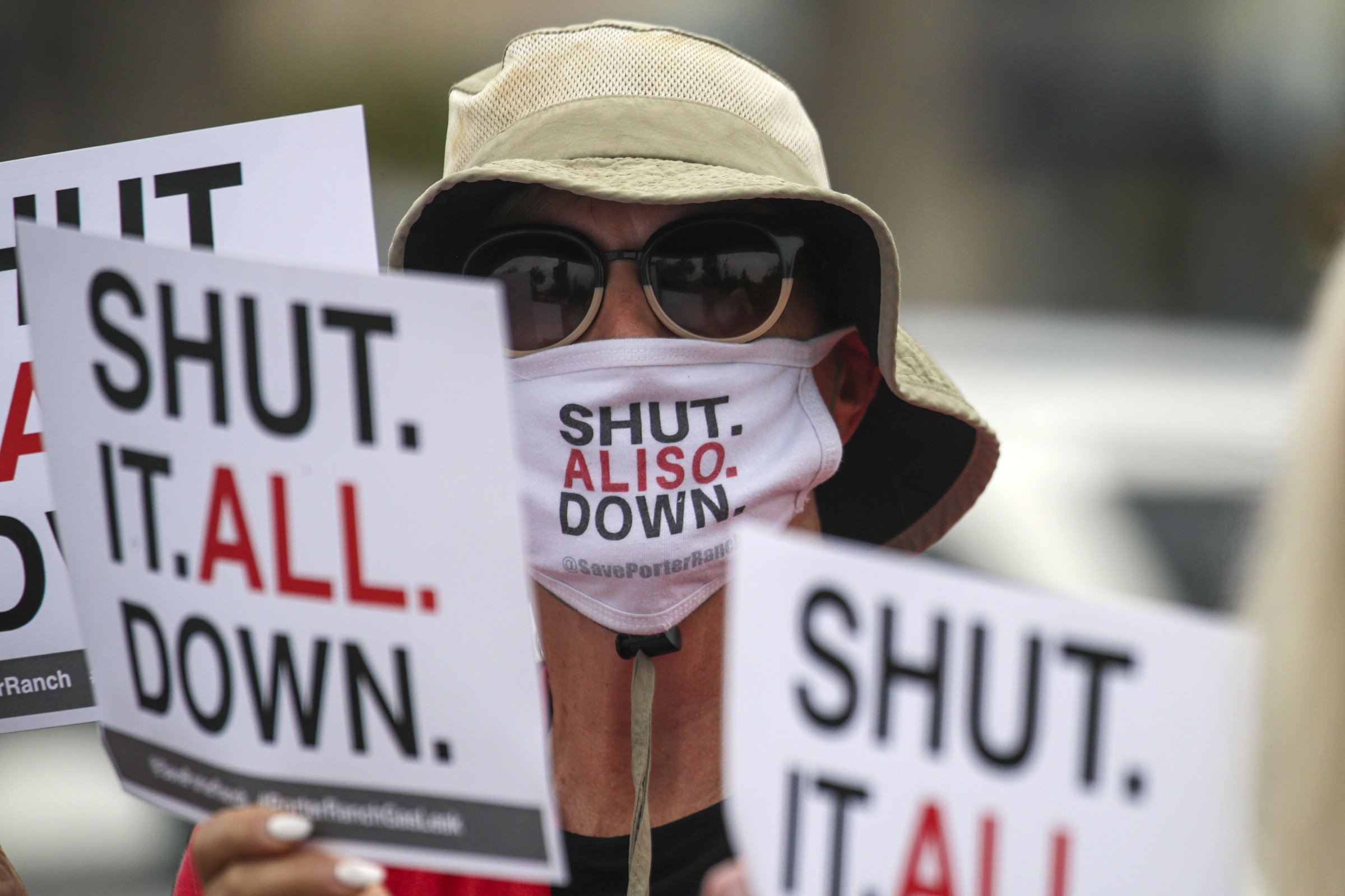 Person wearing a mask with &quot;Shut Aliso Down&quot; on it and signs saying &quot;Shut It All Down&quot;