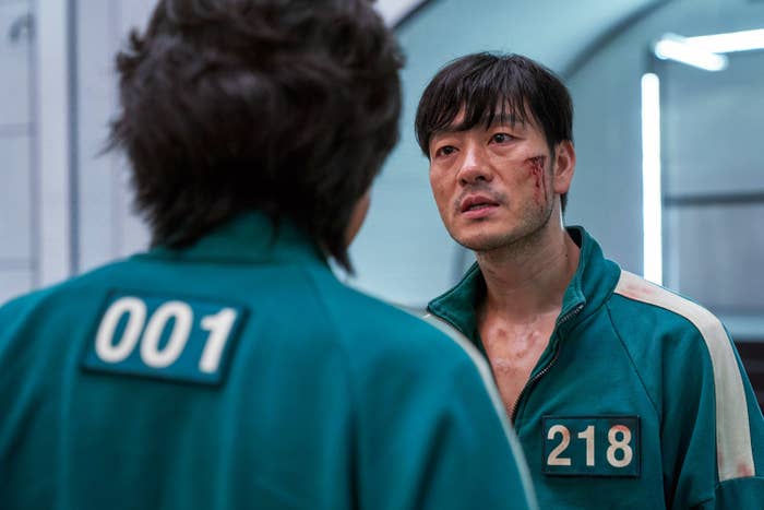 A screengrab from Squid Game shows two people in matching uniforms, one labeled "001" and the other "218" facing each other, the one labeled "218" with a bloody cut on his cheek