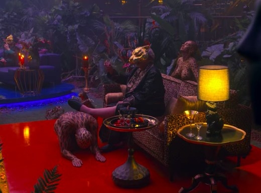 One of the VIPs sits on a couch with his foot propped up on a kneeling woman in body paint; another body-painted woman stands behind the couch with her eyes closed