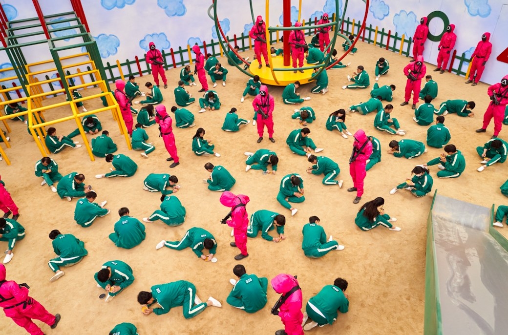The contestants kneel on the ground in an oversized playground setting with guards watching over them