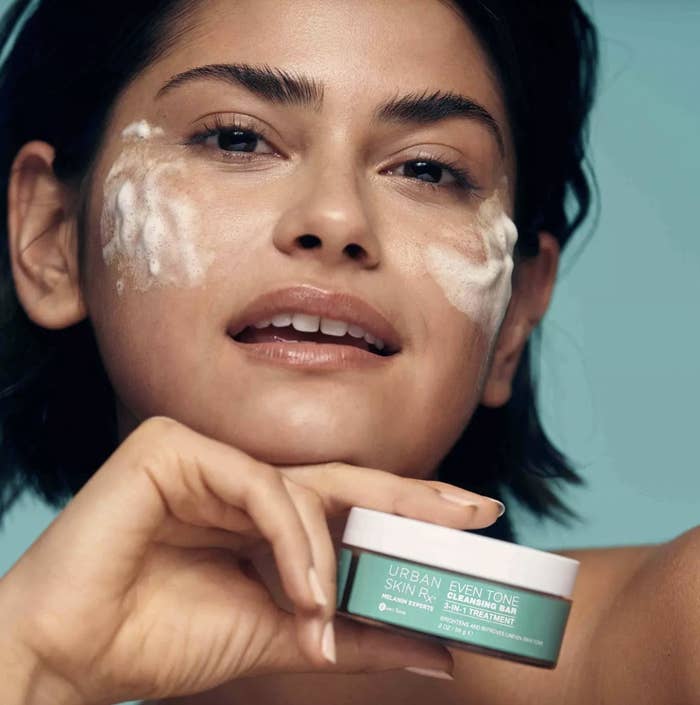 A woman holding a jar of facial cleanser with product on her face