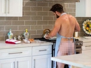 The man stands at the stove wearing nothing but an apron