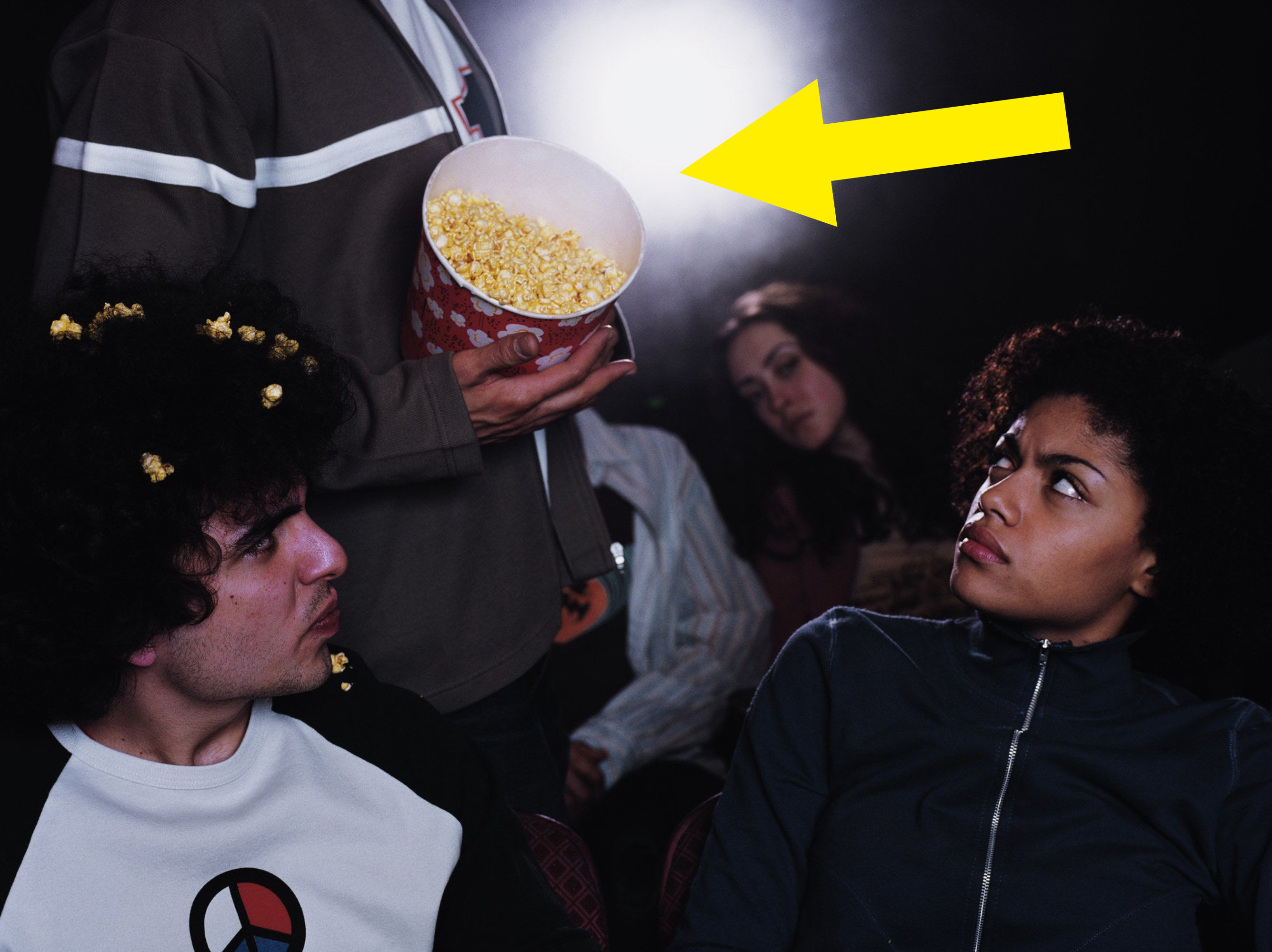 At a movie theater, two people look annoyed as someone passes by and spills popcorn on them
