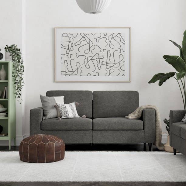 the gray sofa with white and gray accent pillows on it, a brown ottoman in front of it, and a framed piece of artwork hanging on the wall behind it