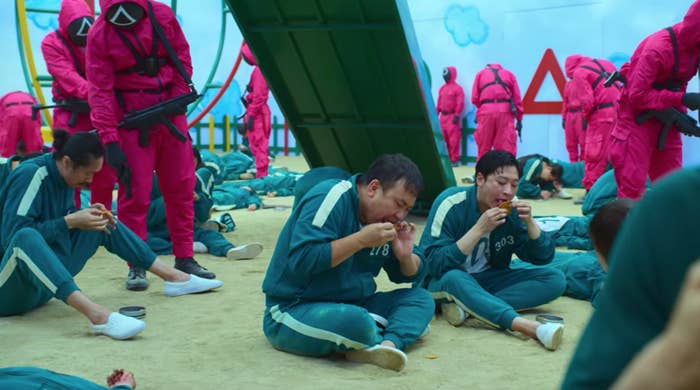 Masked guards with guns stand over contestants who are trying to take a shape out of a tin of honeycomb