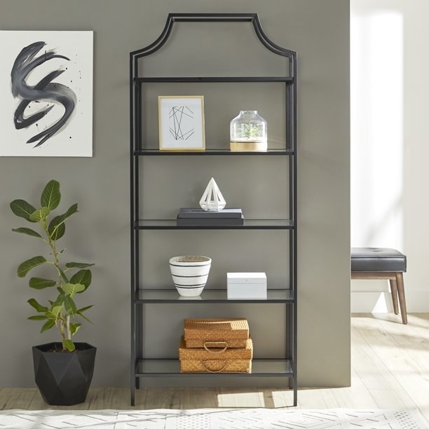 the open bookshelf in black with tan boxes on the bottom shelf, a white vase and white box on the second shelf, books and a triangle decor item on the third shelf, and a framed picture and plant on the fourth shelf