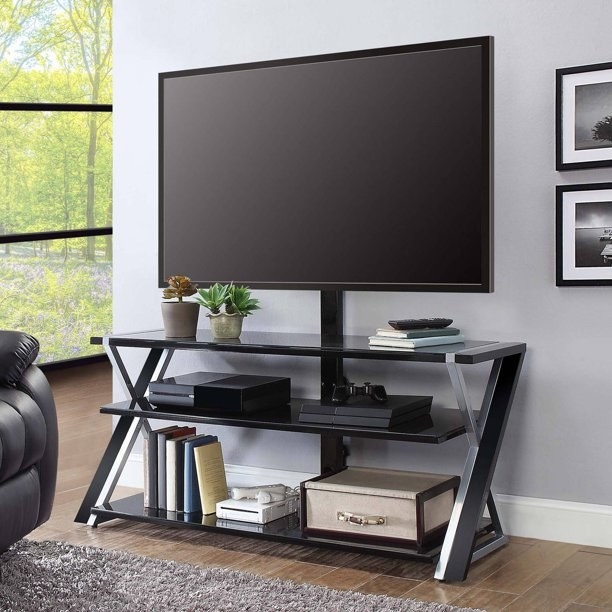 the silver and gray tv stand with criss-crossed legs and open shelves. On the bottom shelf are books, a Wii, and a chest. On the top shelf is a dvd player and a game console. the tv sits on the very top counter with plants and a book