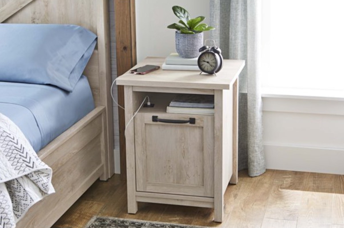 the light brown nightstand with a clock, books, plants, and charging phone on top plugged into the USB port in the middle shelf