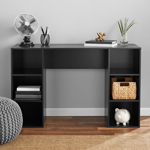 the black desk with a fan, books, pencil holder, and plants on top and books, a basket, and a piggy bank in the cubbies