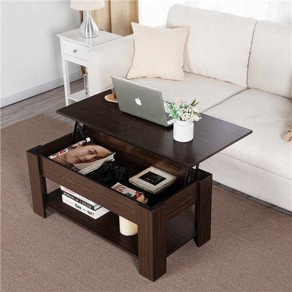 the dark brown coffee table in front of a white couch with the top shelf elevated and a laptop and plant on it with magazines and books in the inside and bottom shelf