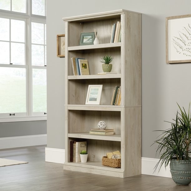 the light brown bookcase with books, plants, picture frames, and baskets in the shelves.