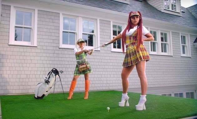 Jesy being handed a golf club while wearing a polo and plaid vest and skirt