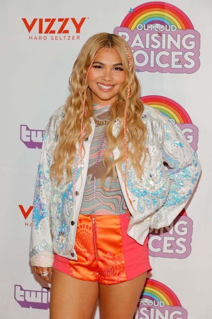 Hayley Kiyoko poses on a red carpet in brightly-colored shorts, a blouse, and a sequined jacket
