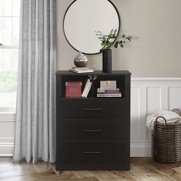 the black dresser with three drawers and an open top section with books and a box in it.