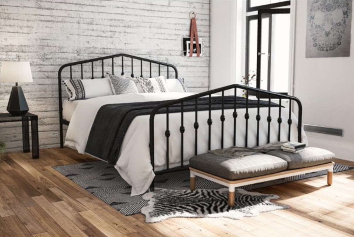 the brown metal headboard with white and black bedding, a bench in front if it and a nightstand with a lamp next it it