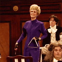 a gif of Kristen Wiig high kicking while wearing a one piece pantsuit and carrying a musical triangle