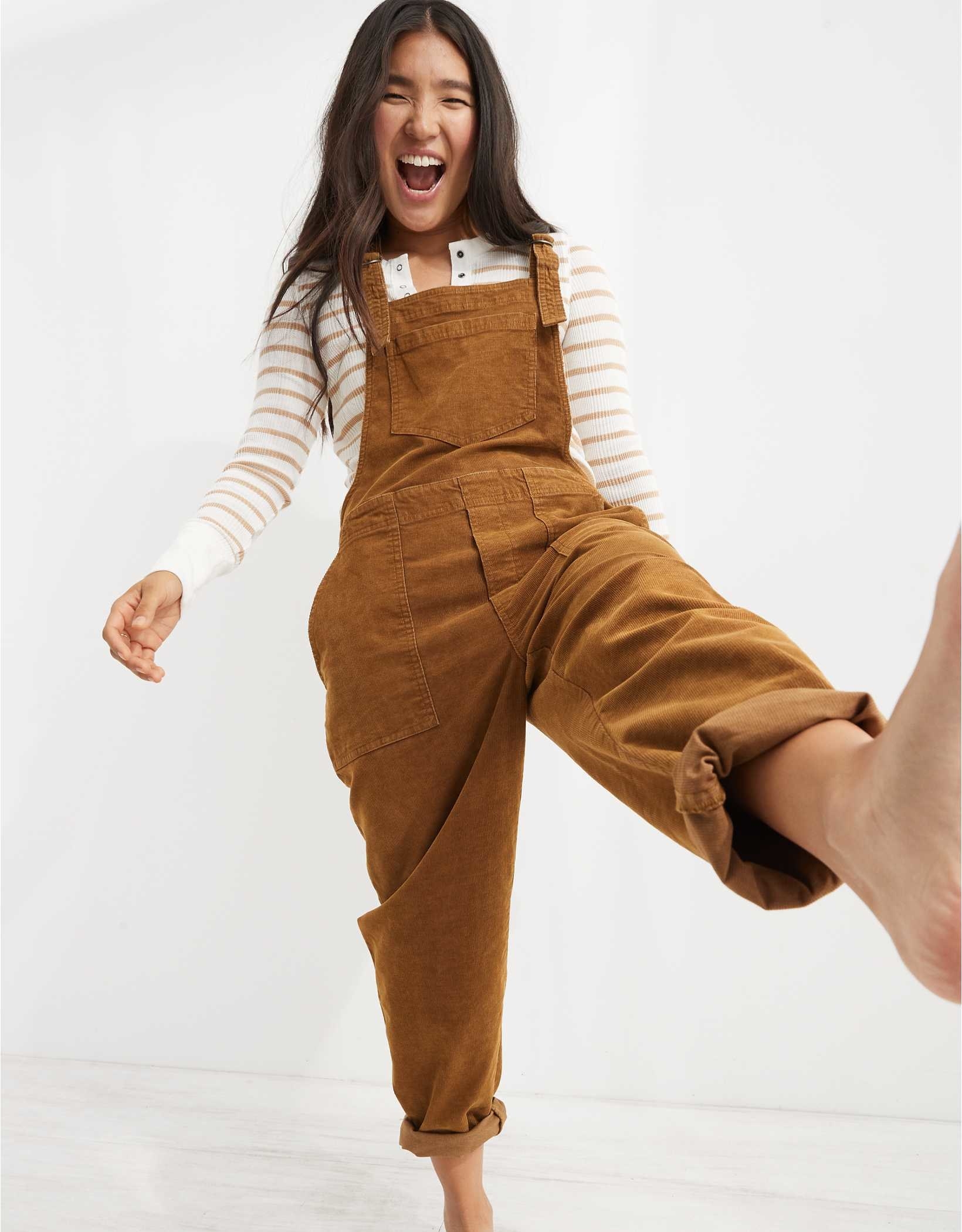 model wearing the tan overalls with a tan and white striped long sleeve top underneath and one leg up in a kick