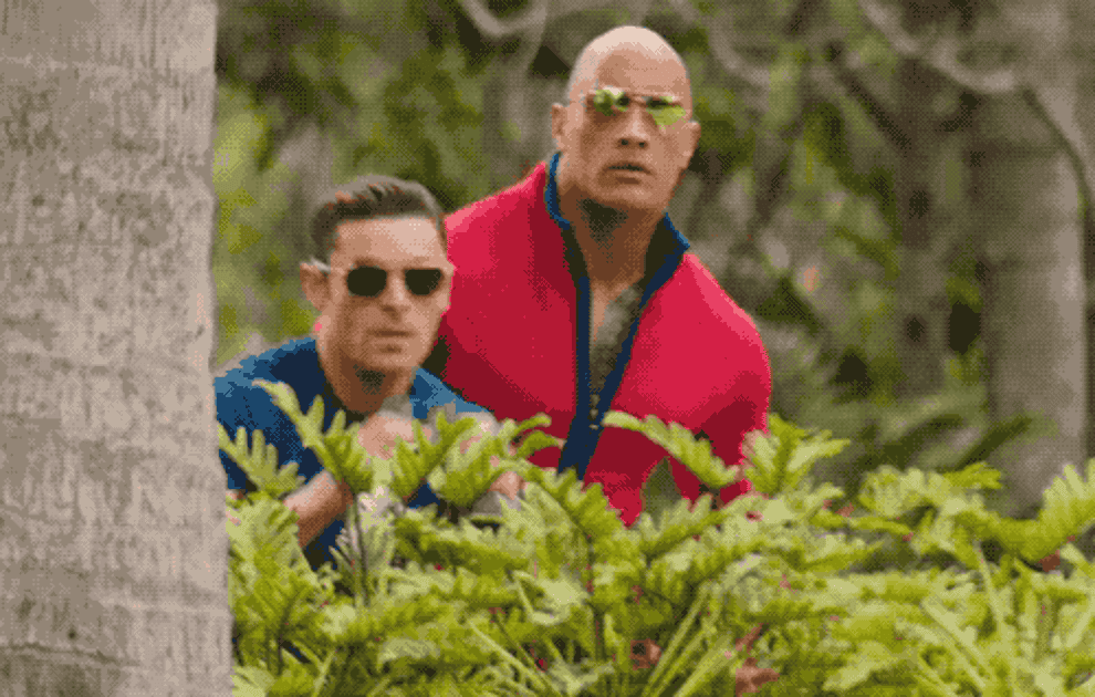a gif of Zac Efron and The Rock peering around a tree with binoculars in the &quot;Baywatch&quot; movie