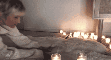 kristen wiig wearing a cozy sweater sliding across a blanket with candles around her