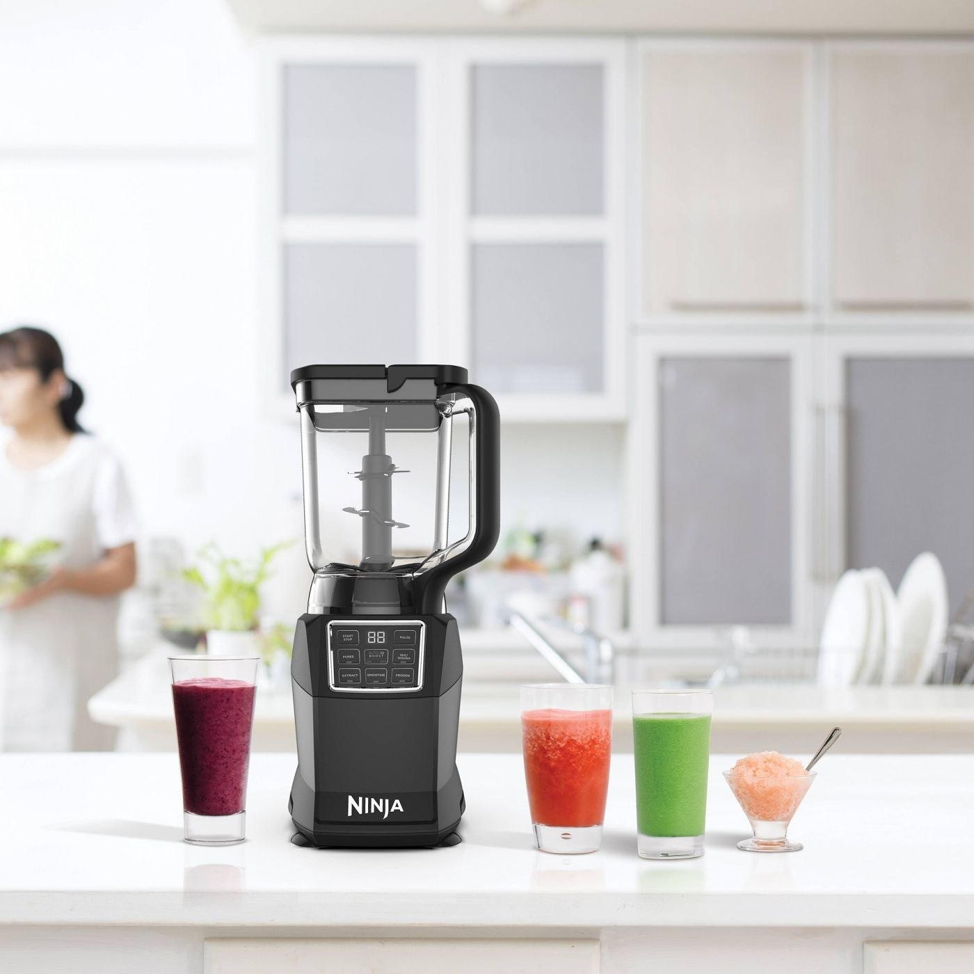 the blender on a counter with several smoothies and a model in the background