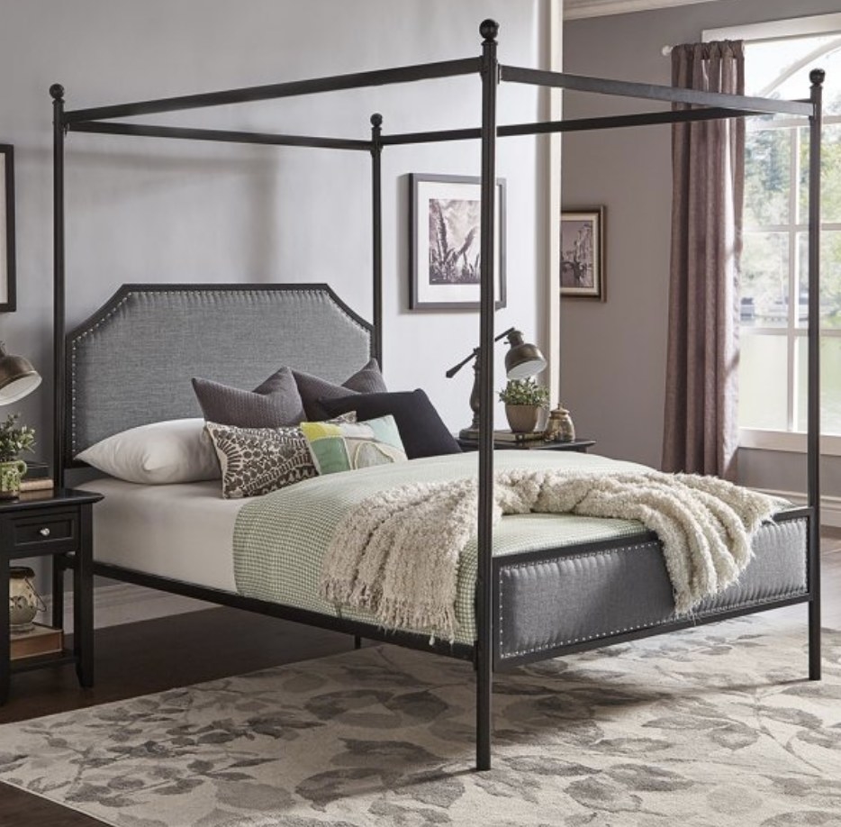 A black canopy bed with grey upholstered headboard and footboard with nailhead trim