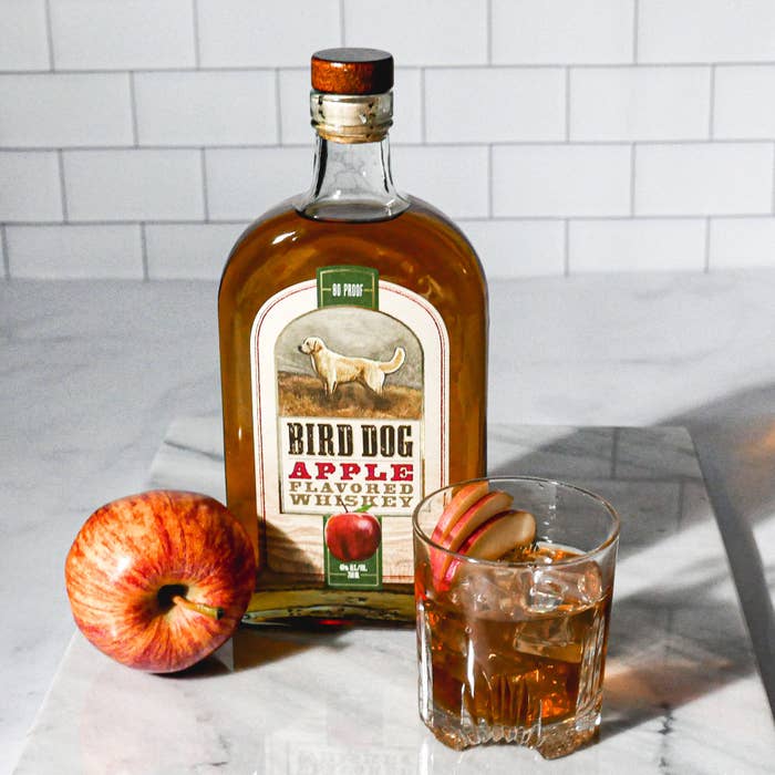 Bird Dog Apple Flavored Whiskey on a county next to an apple and a cocktail in a glass