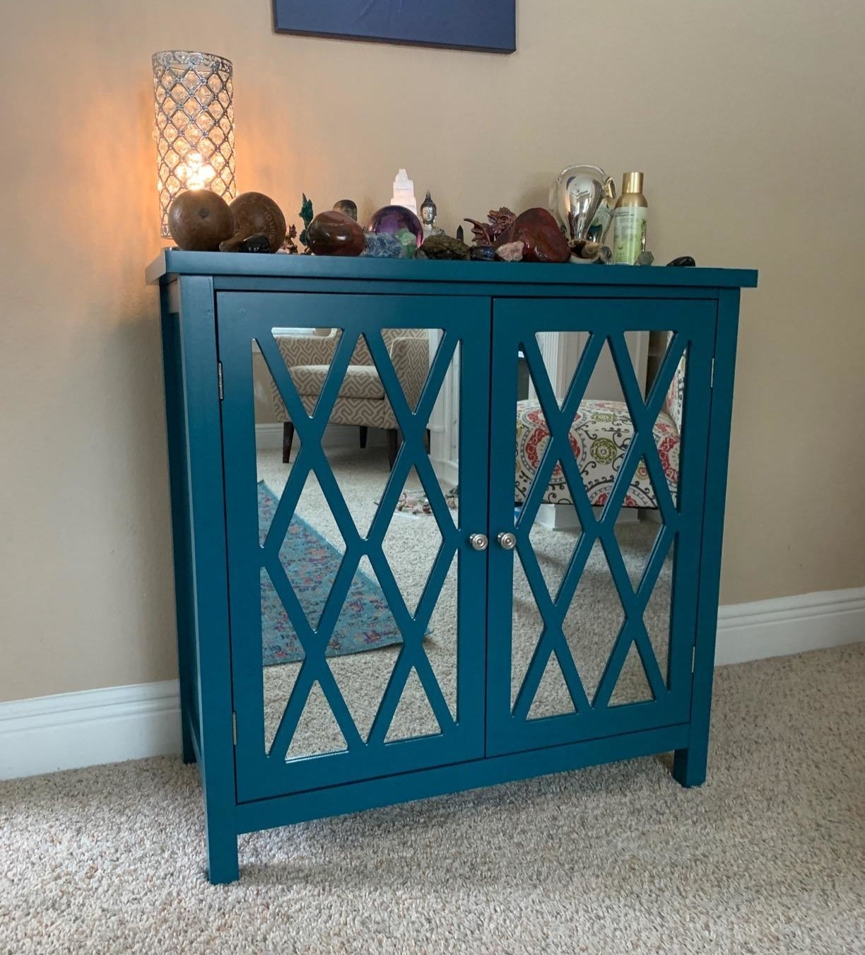 A teal cabinet with crisscross patterned doors, decorative items on top, in a room next to a lit lamp