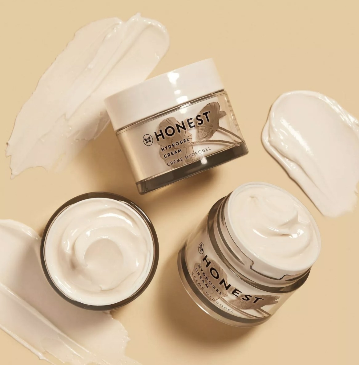 There are three &quot;HONEST&quot; cream jars placed in various positions against a cream background with various swatches in the background