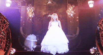 Samantha walks down a staircase in a white gown and masquerade mask