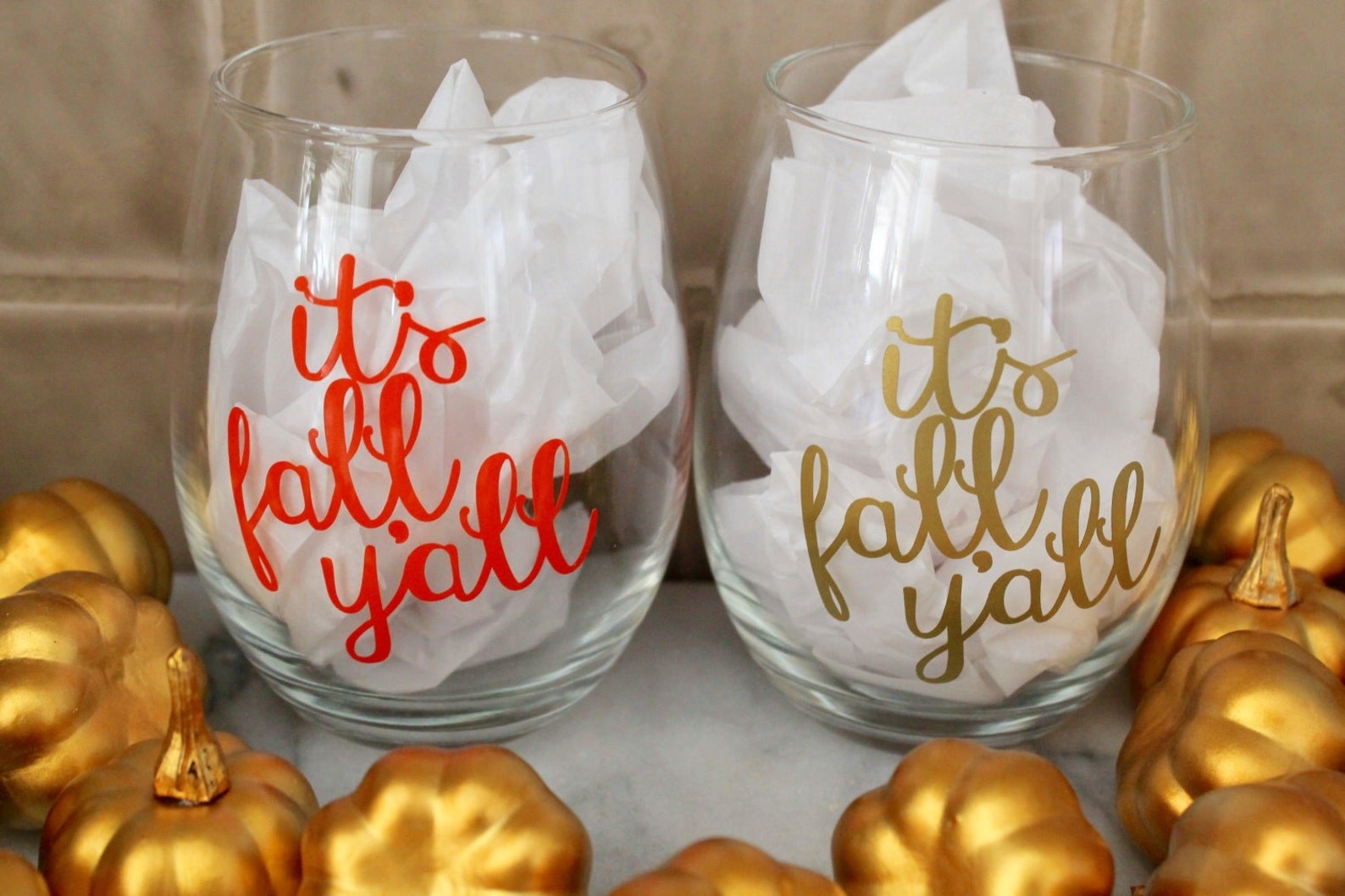 Two clear stemless wine glasses with &quot;it&#x27;s fall, y&#x27;all&quot; written, one in orange and the other in gold