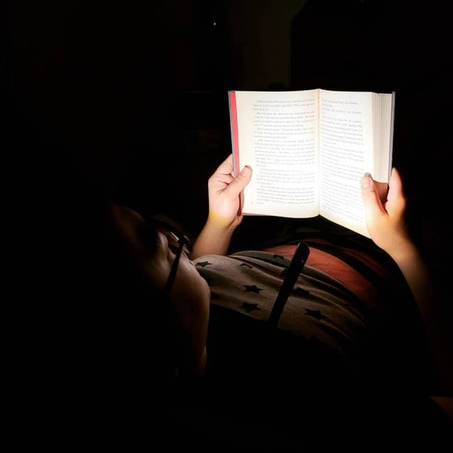 reviewer laying in an all dark room with the reading light around their neck illuminating the book in front of them