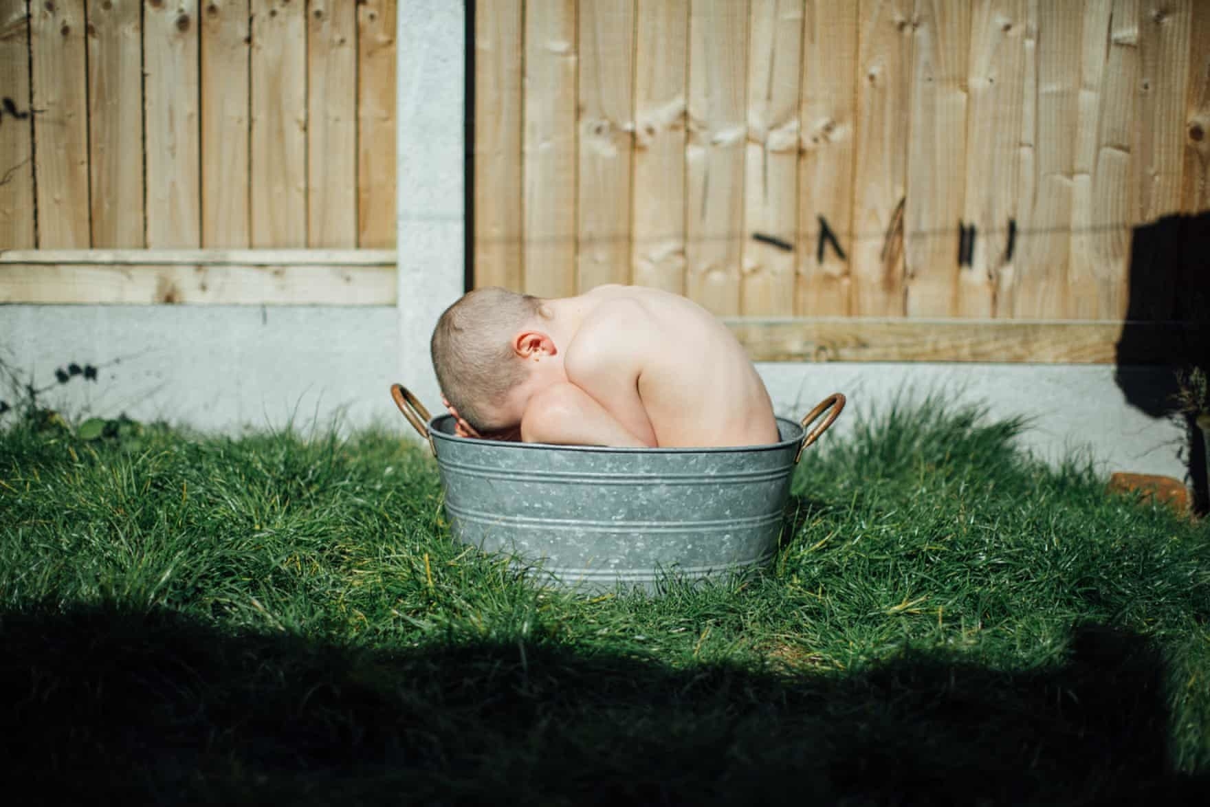 A young boy with close-cropped hair shits in a pail on the grass in front of a wooden fence